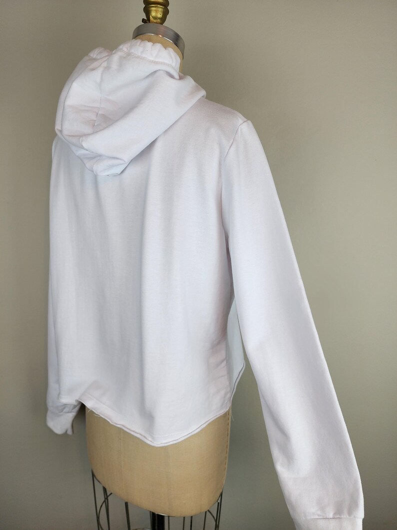Puede incluir: Una sudadera blanca lisa de manga larga con capucha sin cordones, mangas largas con pu&ntilde;os acanalados y un dobladillo recto. La prenda se muestra sobre un maniqu&iacute;, exhibiendo la vista posterior de la tela blanca s&oacute;lida.