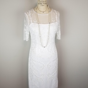 May include: A white beaded dress with a sheer overlay and a scoop neckline. The dress has a fitted silhouette and is adorned with delicate floral embroidery. The dress is displayed on a mannequin.