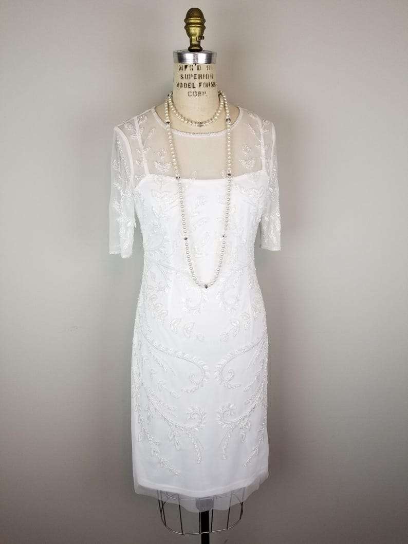 May include: A white beaded dress with a sheer overlay and a scoop neckline. The dress has a fitted silhouette and is adorned with delicate floral embroidery. The dress is displayed on a mannequin.