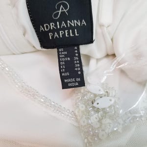 May include: White dress tag with black text that reads "Adrianna Papell" and size information in US, Canadian, UK, EU/FR, German, Spanish, Italian, and Australian sizes. The tag also reads "Made in India".