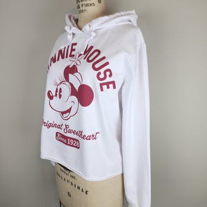 Puede incluir: Una sudadera blanca de manga larga con capucha y un gr&aacute;fico granate de Minnie Mouse. El texto dice Minnie Mouse, Original Sweetheart y Since 1928. La sudadera tiene una capucha con cord&oacute;n y un dobladillo sin rematar en la cintura.