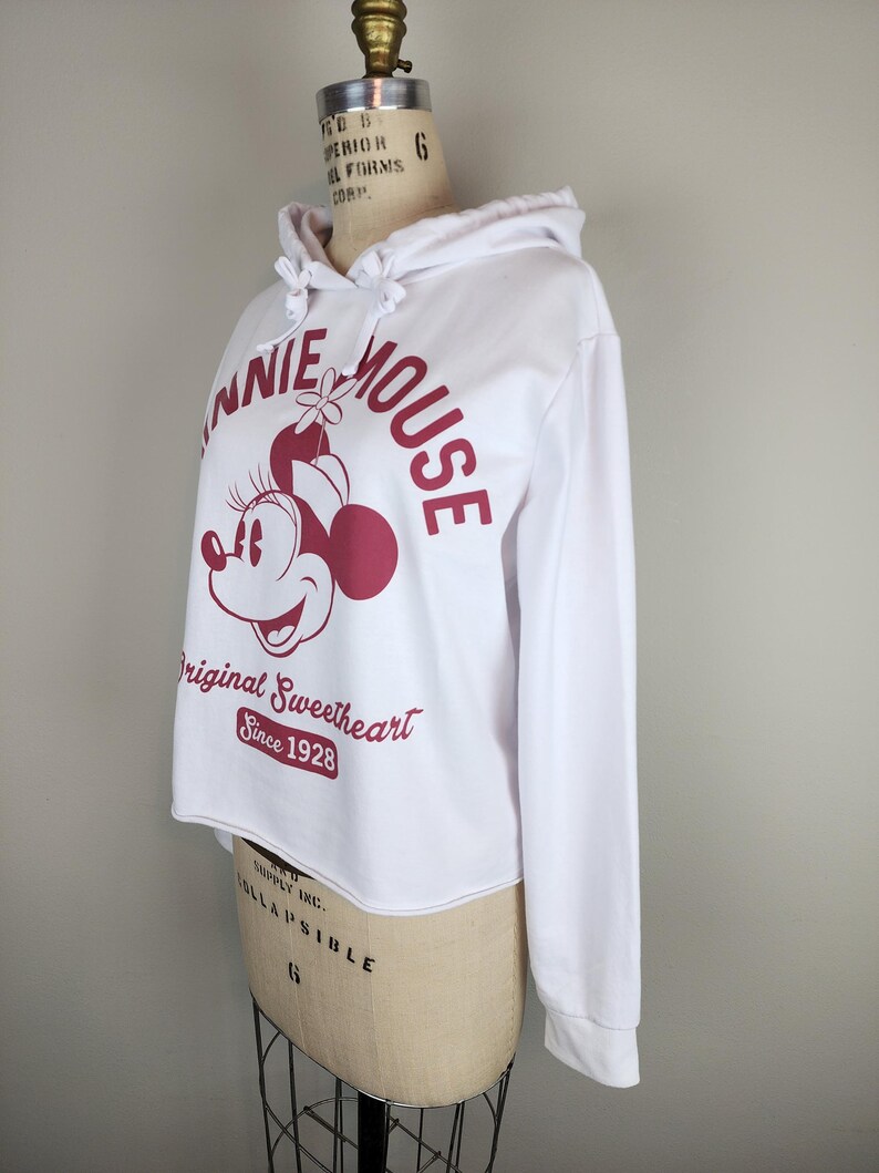 Puede incluir: Una sudadera blanca de manga larga con capucha y un gr&aacute;fico granate de Minnie Mouse. El texto dice Minnie Mouse, Original Sweetheart y Since 1928. La sudadera tiene una capucha con cord&oacute;n y un dobladillo sin rematar en la cintura.