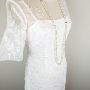 May include: A white beaded dress with a sheer overlay and a beaded neckline. The dress has a fitted silhouette and a short sleeve. The dress is paired with a pearl necklace.