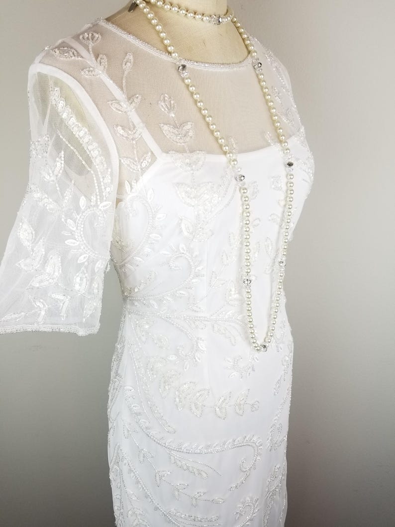 May include: A white beaded dress with a sheer overlay and a beaded neckline. The dress has a fitted silhouette and a short sleeve. The dress is paired with a pearl necklace.