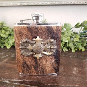 Könnte beinhalten: Eine Edelstahl-Flachmann mit braun-schwarzem Kuhfell-Außenmaterial. Die Flasche hat ein dekoratives Metallabzeichen mit Kreuz- und Wappenmuster. Die Oberseite der Flasche ist silberfarben.