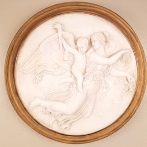 Op de afbeelding: Een ronde, witte reliëfsculptuur van een vrouw en een kind met vleugels, geplaatst in een goudkleurige houten lijst. De figuren dragen vloeiende kleding, waarbij het kind omhoog reikt. Het kunstwerk is een klassieke afbeelding, mogelijk een engel of een mythologische scène.