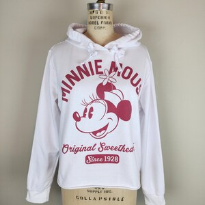 Puede incluir: Una sudadera blanca de manga larga con capucha y un gr&aacute;fico rosa oscuro de Minnie Mouse. El texto dice "MINNIE MOUSE", "Original Sweetheart" y "Since 1928" en color rosa oscuro. La sudadera incluye una capucha con cord&oacute;n y mangas largas.