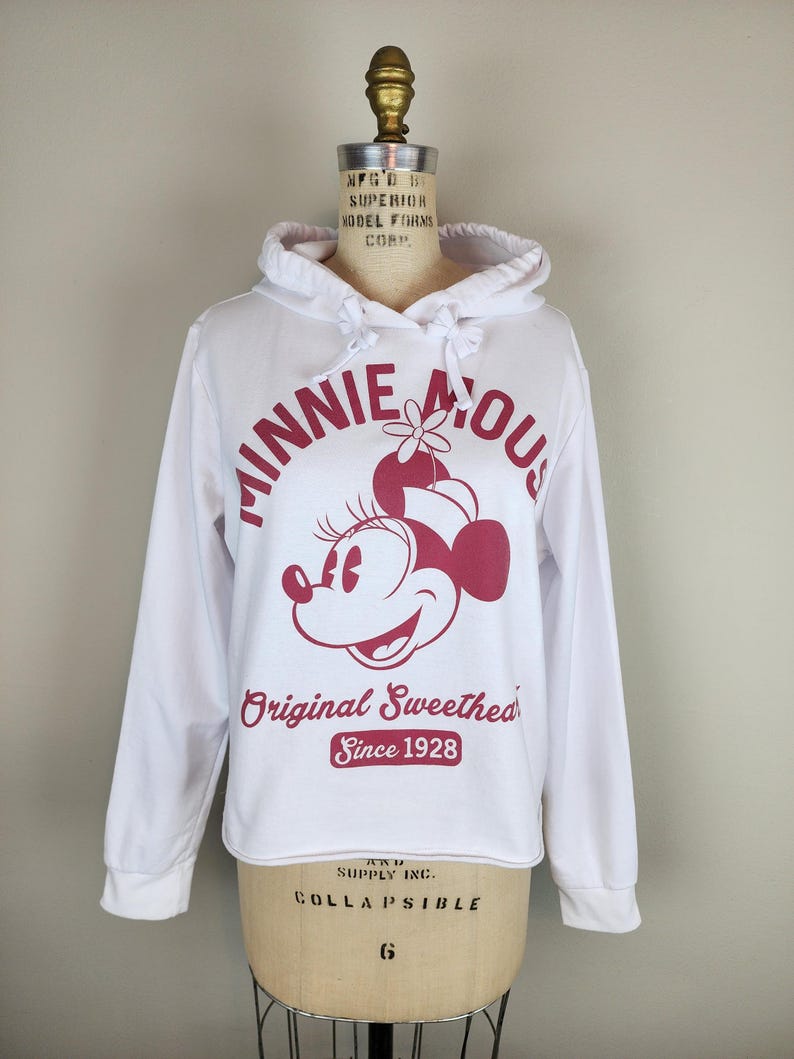 Puede incluir: Una sudadera blanca de manga larga con capucha y un gr&aacute;fico rosa oscuro de Minnie Mouse. El texto dice "MINNIE MOUSE", "Original Sweetheart" y "Since 1928" en color rosa oscuro. La sudadera incluye una capucha con cord&oacute;n y mangas largas.