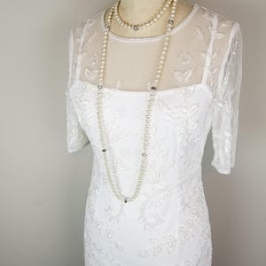 May include: A white beaded dress with a sheer overlay and a pearl necklace. The dress has a fitted bodice and a flowing skirt.