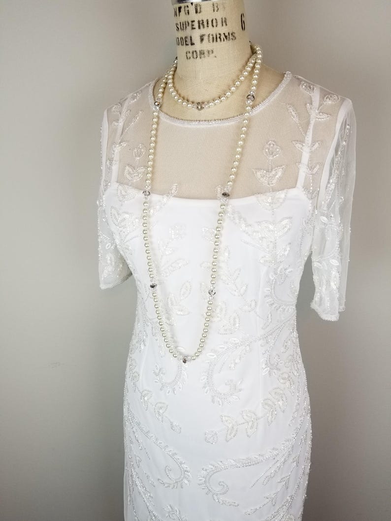 May include: A white beaded dress with a sheer overlay and a pearl necklace. The dress has a fitted bodice and a flowing skirt.