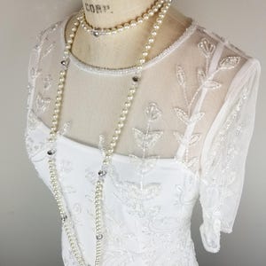 May include: A white beaded dress with a sheer overlay and a pearl and crystal necklace.