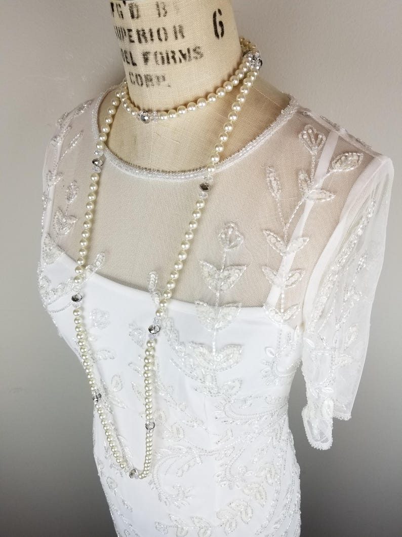 May include: A white beaded dress with a sheer overlay and a pearl and crystal necklace.