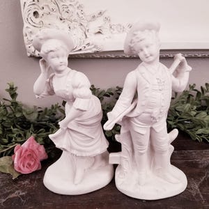 Puede incluir: Dos figuritas de porcelana blanca de un niño y una niña. El niño sostiene un palo y la niña lleva un vestido y un sombrero. Las figuritas están de pie sobre una base blanca.
