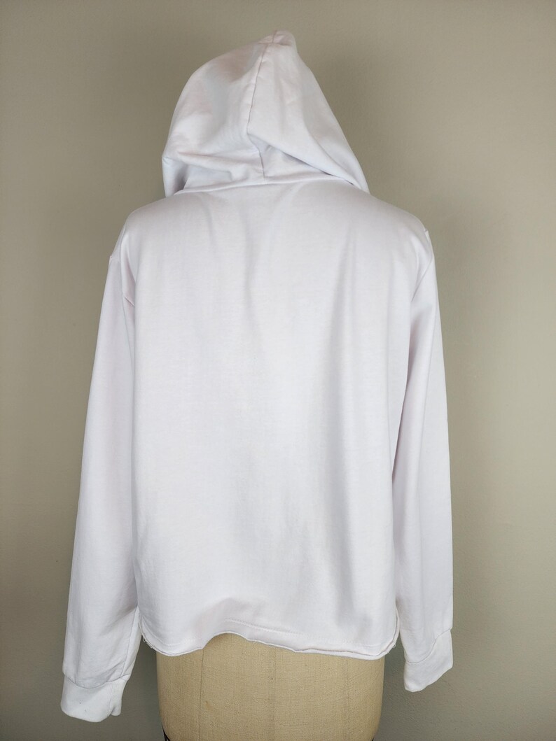 Puede incluir: Una sudadera con capucha blanca de manga larga, con una capucha sin cordones, mangas largas con pu&ntilde;os acanalados y un dobladillo recto. La prenda est&aacute; confeccionada con un tejido liso y sencillo, y se muestra desde la vista posterior.