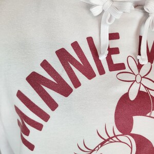 Puede incluir: Una sudadera con capucha blanca que presenta un estampado gr&aacute;fico en rojo oscuro del texto MINNIE y una ilustraci&oacute;n parcial de un personaje con una flor. La sudadera incluye un cord&oacute;n blanco a juego con extremos anudados en el cuello.