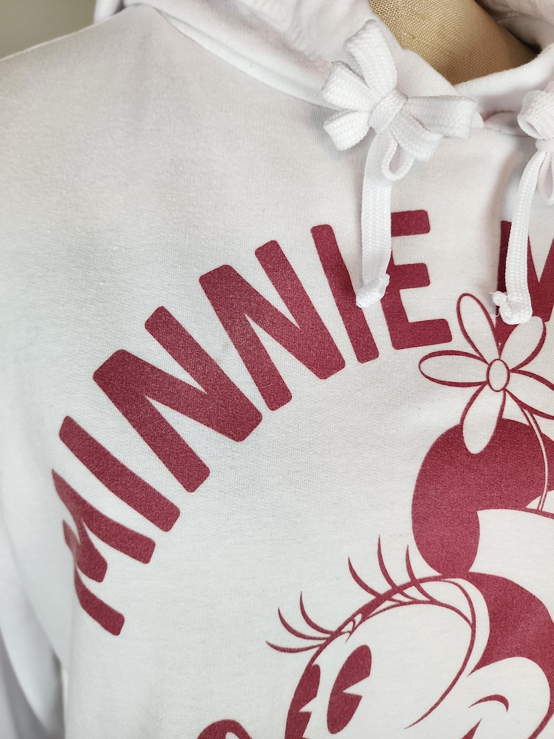 Puede incluir: Una sudadera con capucha blanca que presenta un estampado gr&aacute;fico en rojo oscuro del texto MINNIE y una ilustraci&oacute;n parcial de un personaje con una flor. La sudadera incluye un cord&oacute;n blanco a juego con extremos anudados en el cuello.