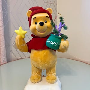 May include: A plush Winnie the Pooh figure wearing a red knit hat and red shirt, standing on a white base. The figure holds a yellow star in one hand and a green pot labeled HUNNY with a small artificial pine tree in the other hand.