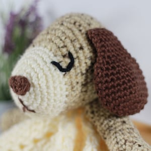 May include: A crocheted stuffed dog toy with brown ears, a brown nose, and black embroidered eyes. The dog is lying on a crocheted blanket.