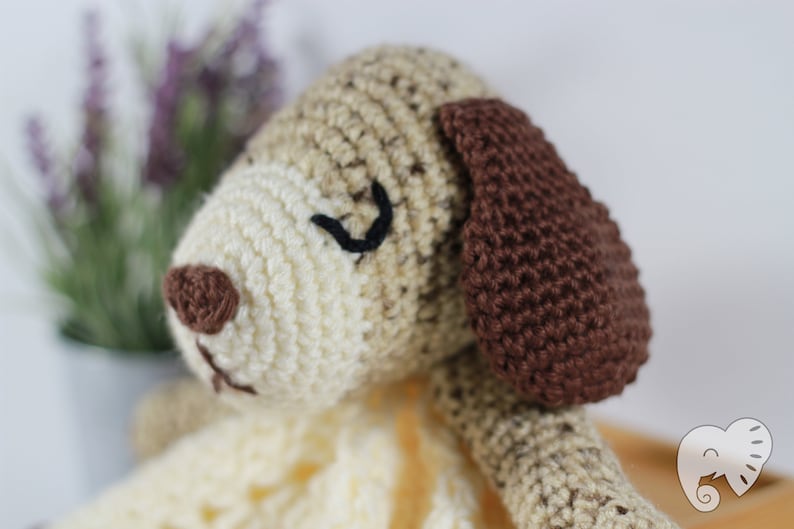 May include: A crocheted stuffed dog toy with brown ears, a brown nose, and black embroidered eyes. The dog is lying on a crocheted blanket.