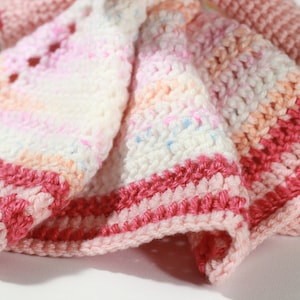 May include: A crocheted baby blanket with pink, white, and peach stripes. The blanket has a delicate, lacy pattern and a pink border.