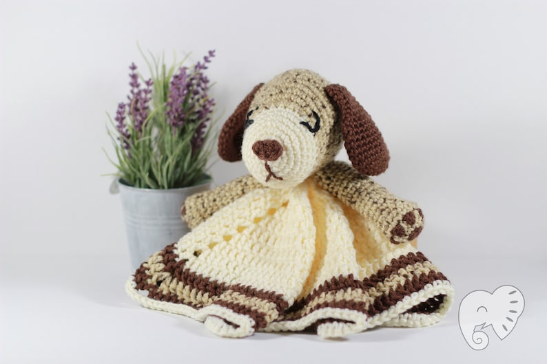 May include: A crocheted dog-shaped baby blanket with brown and beige stripes. The blanket has a crocheted dog head with brown ears and a brown nose.
