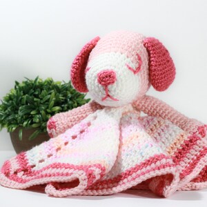 May include: A pink and white crocheted puppy with a blanket attached. The puppy has a white face with pink ears and a pink nose. The blanket is a multi-colored pink and white with a crocheted border.