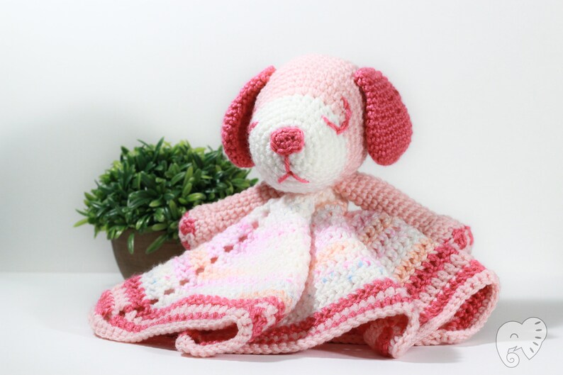 May include: A pink and white crocheted puppy with a blanket attached. The puppy has a white face with pink ears and a pink nose. The blanket is a multi-colored pink and white with a crocheted border.