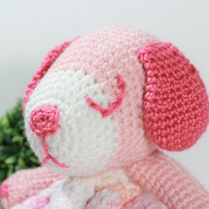 May include: A pink and white crocheted stuffed animal dog with embroidered eyes and a pink nose. The dog is sitting on a white background.