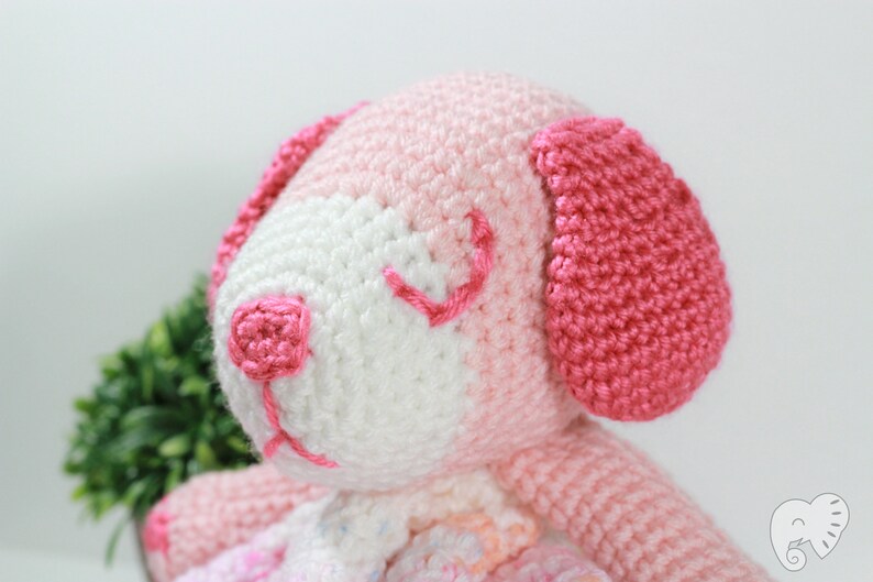 May include: A pink and white crocheted stuffed animal dog with embroidered eyes and a pink nose. The dog is sitting on a white background.