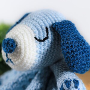 May include: A crocheted blue and white puppy toy with black eyes and a blue nose. The puppy is lying down with its head tilted to the side.