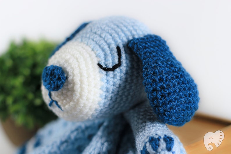May include: A crocheted blue and white puppy toy with black eyes and a blue nose. The puppy is lying down with its head tilted to the side.