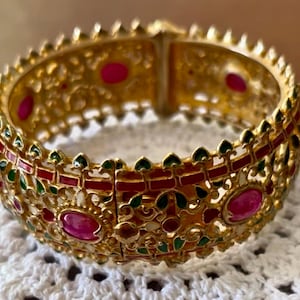 May include: A gold-coloured hinged bangle bracelet with detailed patterns. The bracelet has oval, ruby-red stones and is decorated with red and green enamel. The design incorporates floral and geometric motifs, creating a luxurious appearance.