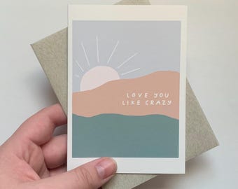 May include: A greeting card with a pastel blue, pink, and green sunset illustration. The text "Love you like crazy" is written in white on the card.