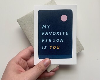 May include: A card with a dark blue background and a pink circle. The text on the card reads "My Favorite Person Is You" in a colorful font.
