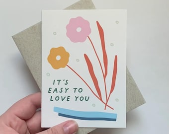 May include: A white card with a green, orange, and pink floral design. The text "It's easy to love you" is written in green and black.