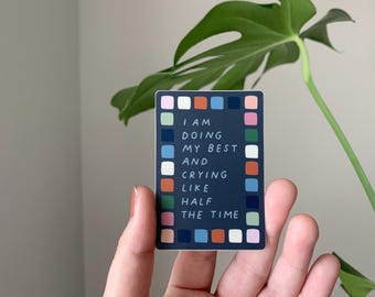 May include: A navy blue sticker with the text "I AM DOING MY BEST AND CRYING LIKE HALF THE TIME." The sticker has a colorful border with various squares. A green plant is in the background.