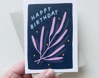 May include: A birthday card with a navy blue background and the words "HAPPY BIRTHDAY" in light blue. The card features a pink and purple botanical illustration with yellow dots. The card is held with a gray envelope.