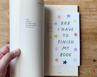 May include: An open book with a bookmark that reads "BRB I HAVE TO FINISH MY BOOK" with colorful star illustrations. The book's pages are cream-colored, and the bookmark is white with green text.