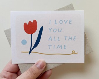 May include: A white card with a red tulip, blue stem, and gold line. The text on the card reads "I love you all the time".
