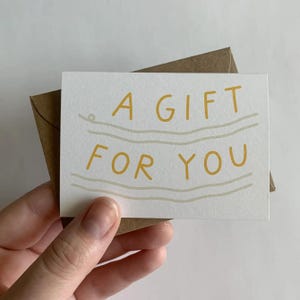 May include: A white card with a brown envelope. The card has yellow text that says "A GIFT FOR YOU" with wavy lines above and below the text.