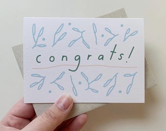 May include: A white card with a green "congrats!" message and blue floral designs. The card has a pink line under the message.