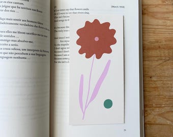 May include: A bookmark with a floral design is placed inside an open book. The bookmark features a large, rust-colored flower with a pink stem and a green dot. The book pages have text in Portuguese and English.