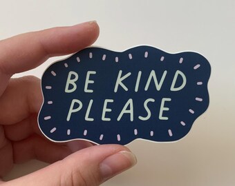 May include: A dark blue sticker with a white outline and pink dots. The sticker has the text "BE KIND PLEASE" in a light green color.