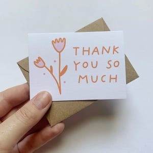 May include: A white card with a pink and orange flower illustration and the text "THANK YOU SO MUCH". The card is in a brown envelope.