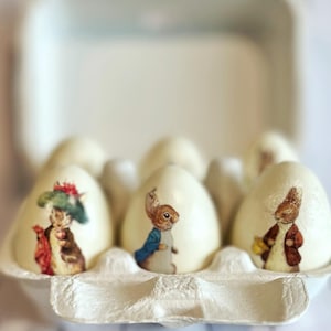 May include: A carton of six white eggs, each decorated with a different illustration of a rabbit in a vintage style. The egg carton is made of recycled paper. The eggs are a light cream color.