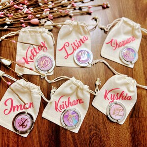 May include: Six small white fabric bags with pink drawstring closures, each containing a silver-toned purse hook with a colorful design and a personalized name in pink lettering. The names are: Kelley, Lisina, Shaquana, Iman, Keishana, and Kyshia.