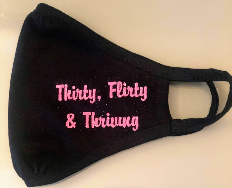 May include: Black face mask with pink glitter lettering that reads "Thirty, flirty & thriving".