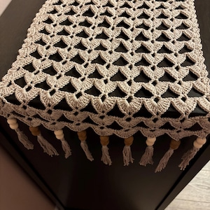 May include: A beige crochet table runner with a geometric pattern. The runner has a scalloped edge with tassels and wooden beads. The handmade table runner is a decorative accent for a dining table or other furniture.