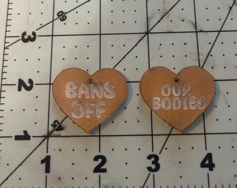 May include: Two light brown heart-shaped wooden charms featuring engraved text. The left charm reads BANS OFF and the right charm reads OUR BODIES. Each charm has a small hole at the top for hanging, positioned on a cutting mat with inch markings.