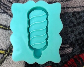 May include: A teal blue silicone mold for making a swirl candy or cake pop. The mold has a wavy edge and a single cavity shaped like a swirl.